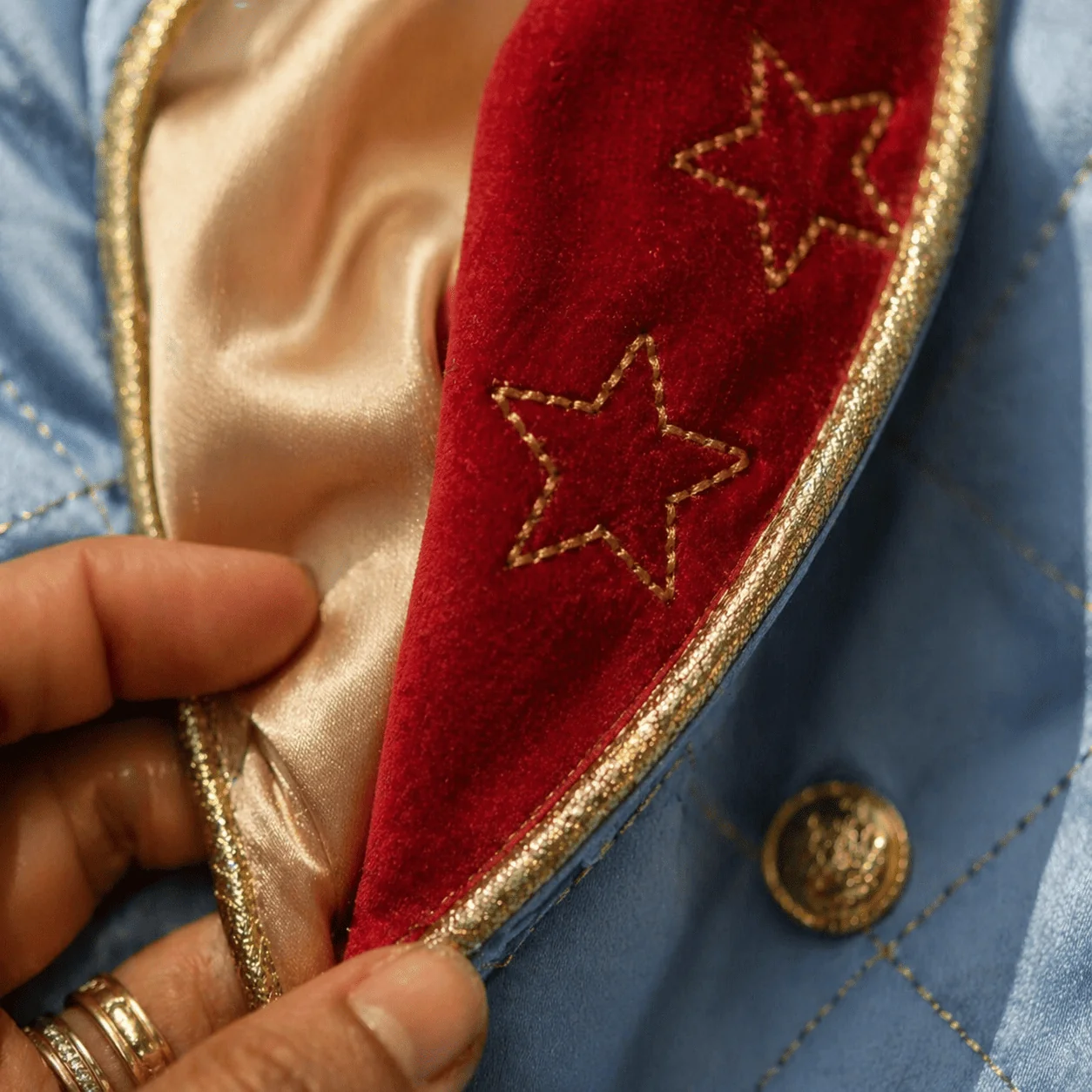 Macro extremo da lapela do casaco Príncipe Luxo aberta parcialmente revelando o forro de seda champanhe por baixo — veludo vermelho com estrela dourada bordada encontra o cetim azul com losangos dourados no pesponto, friso dourado na borda — mão feminina brasileira com aliança discreta segurando a lapela aberta