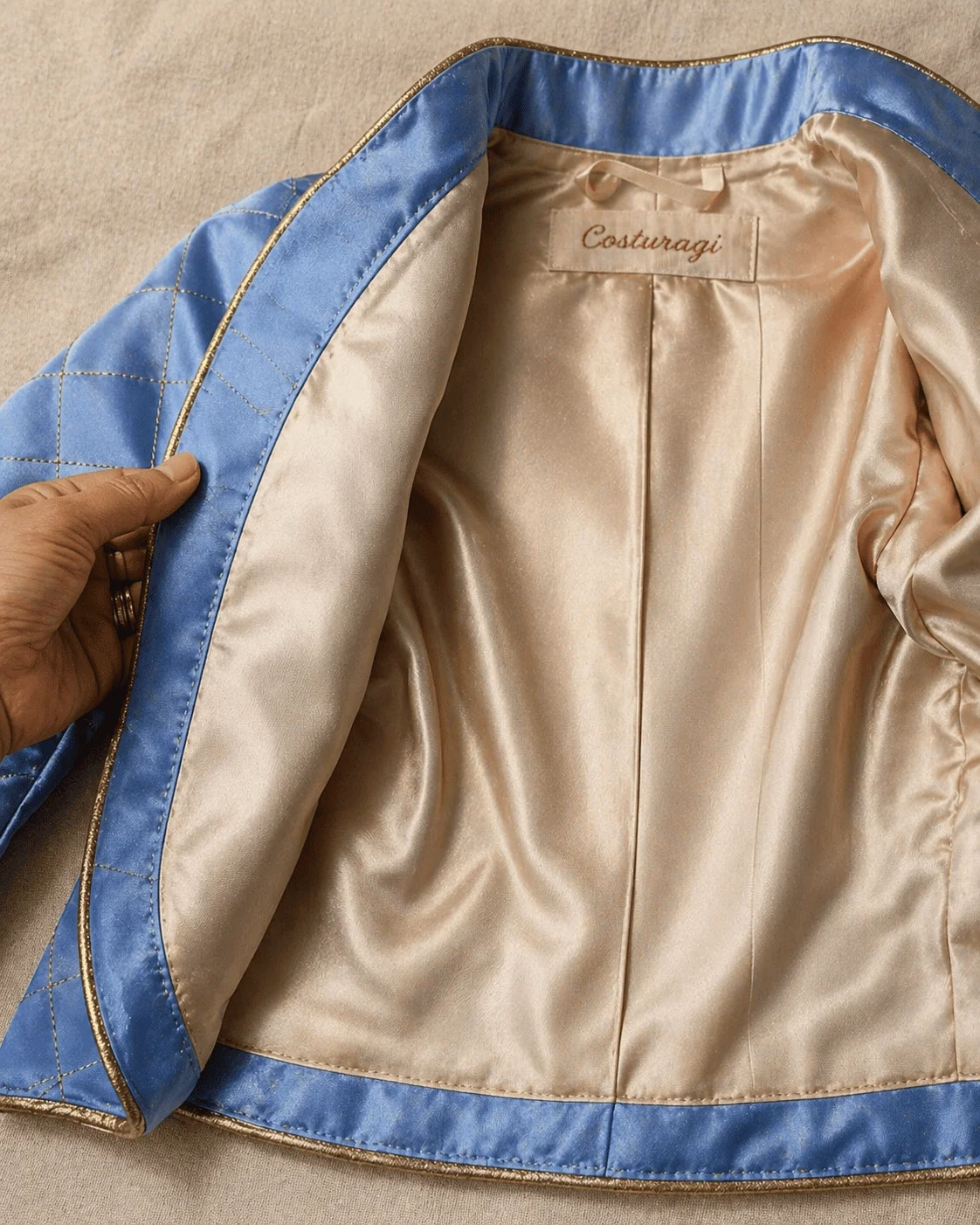 Casaco do Conjunto Príncipe Luxo aberto do avesso sobre superfície de linho cru marfim mostrando o forro de seda champanhe em detalhe — costura invisível à mão na barra, costura interna limpa entre forro e entretela, alcinha de fita cetim no centro das costas com etiqueta Costuragi bordada em linha cobre sobre seda marfim — mão feminina virando uma lapela para revelar o acabamento museu-grade
